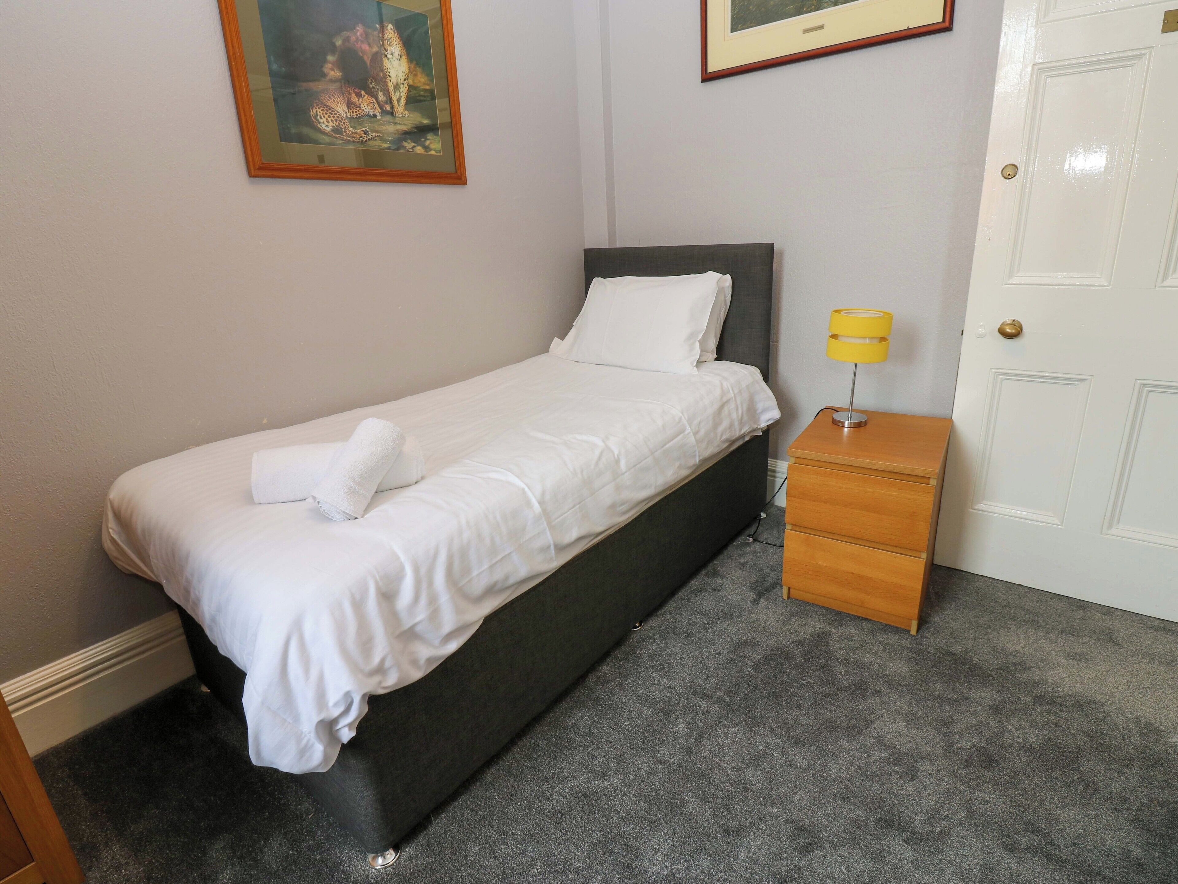 A neatly made bed with white linens and a rolled towel, a bedside table with a yellow lamp, and a door to the right.
