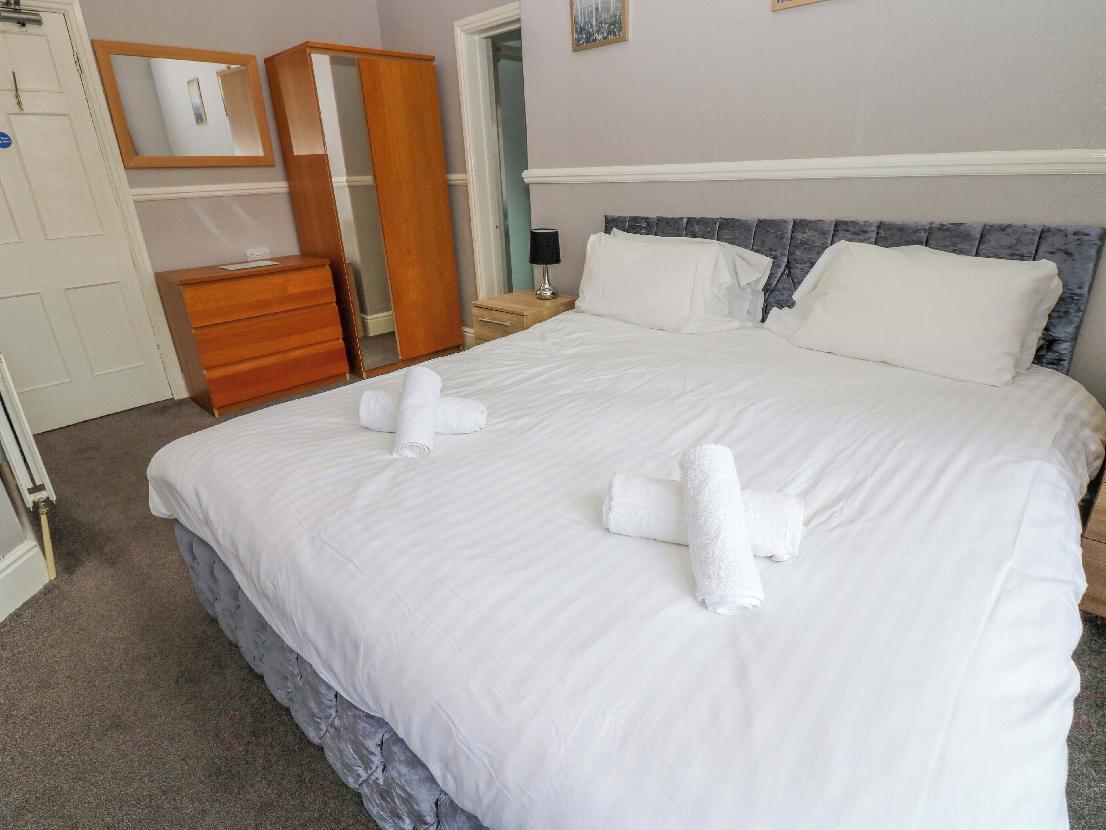 A neatly made bed with white linens and towels, a wooden wardrobe, a mirror, and a nightstand with a lamp.