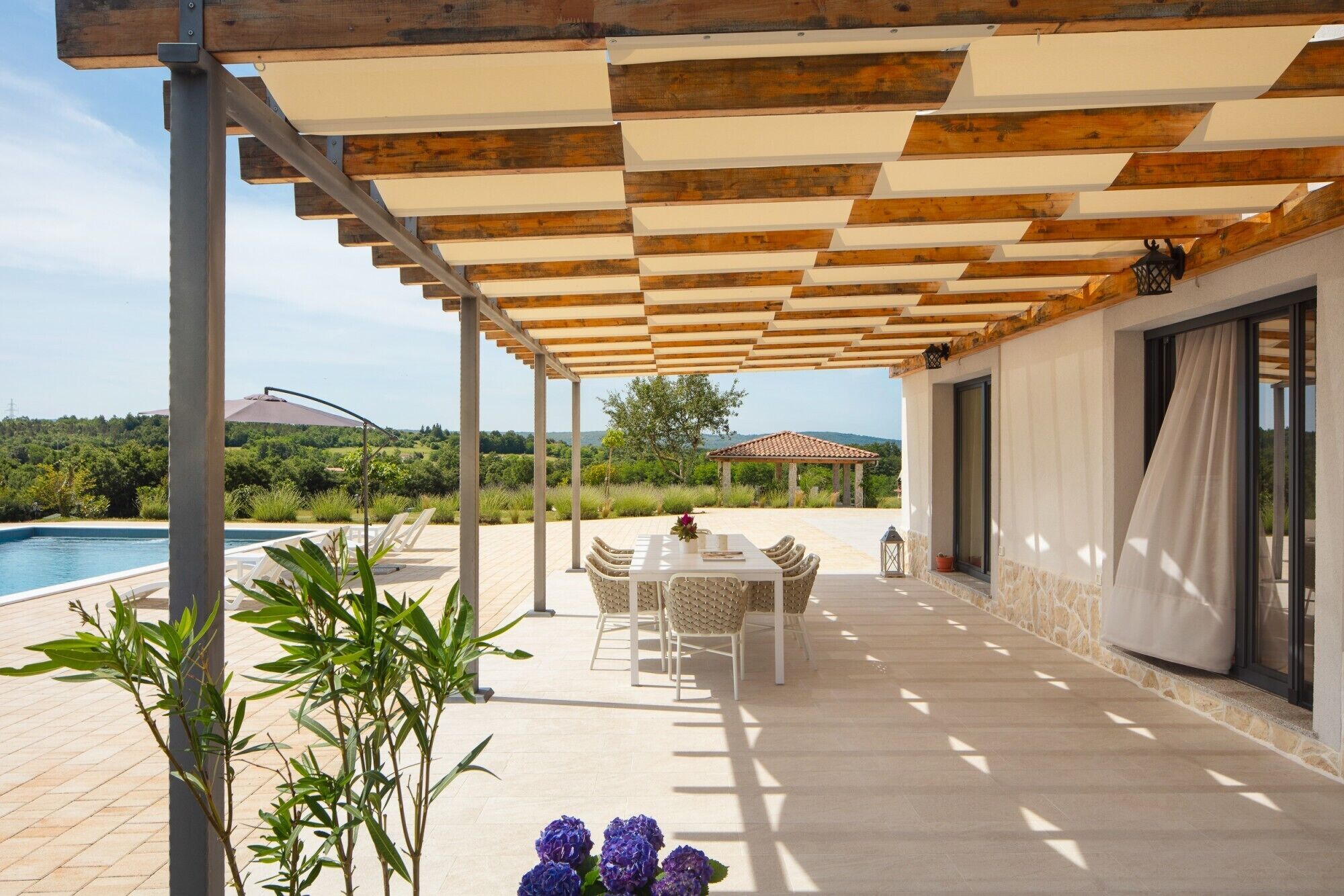 A covered outdoor patio with a pool, a table with chairs, and a view of the surrounding landscape.