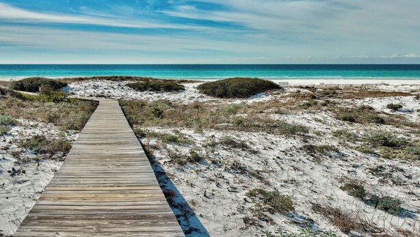 Een houten looppad dat naar een strand leidt, met heldere lucht en rustig water.