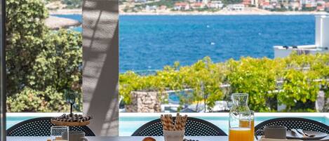 A breakfast table with croissants, berries, and orange juice, overlooking a coastal view with a pool and greenery.
