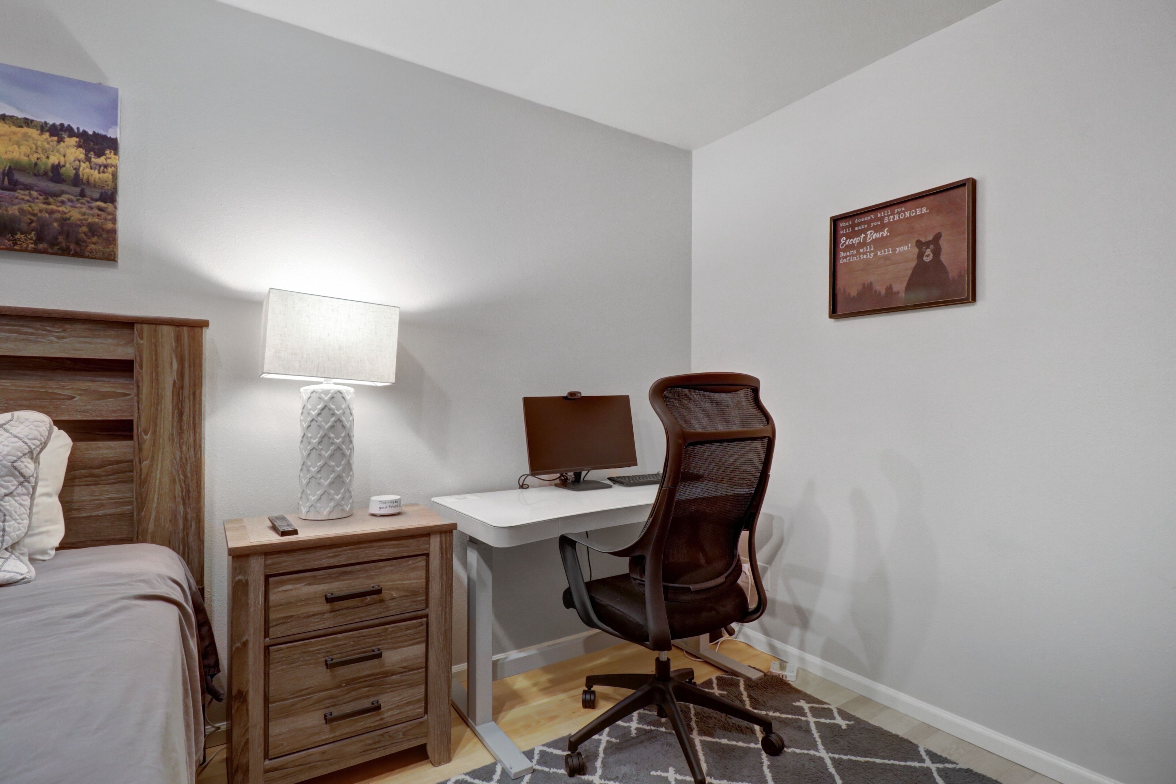 Ett sovrum med en säng, ett skrivbord, en stol, en lampa och ett inramat citat på väggen.