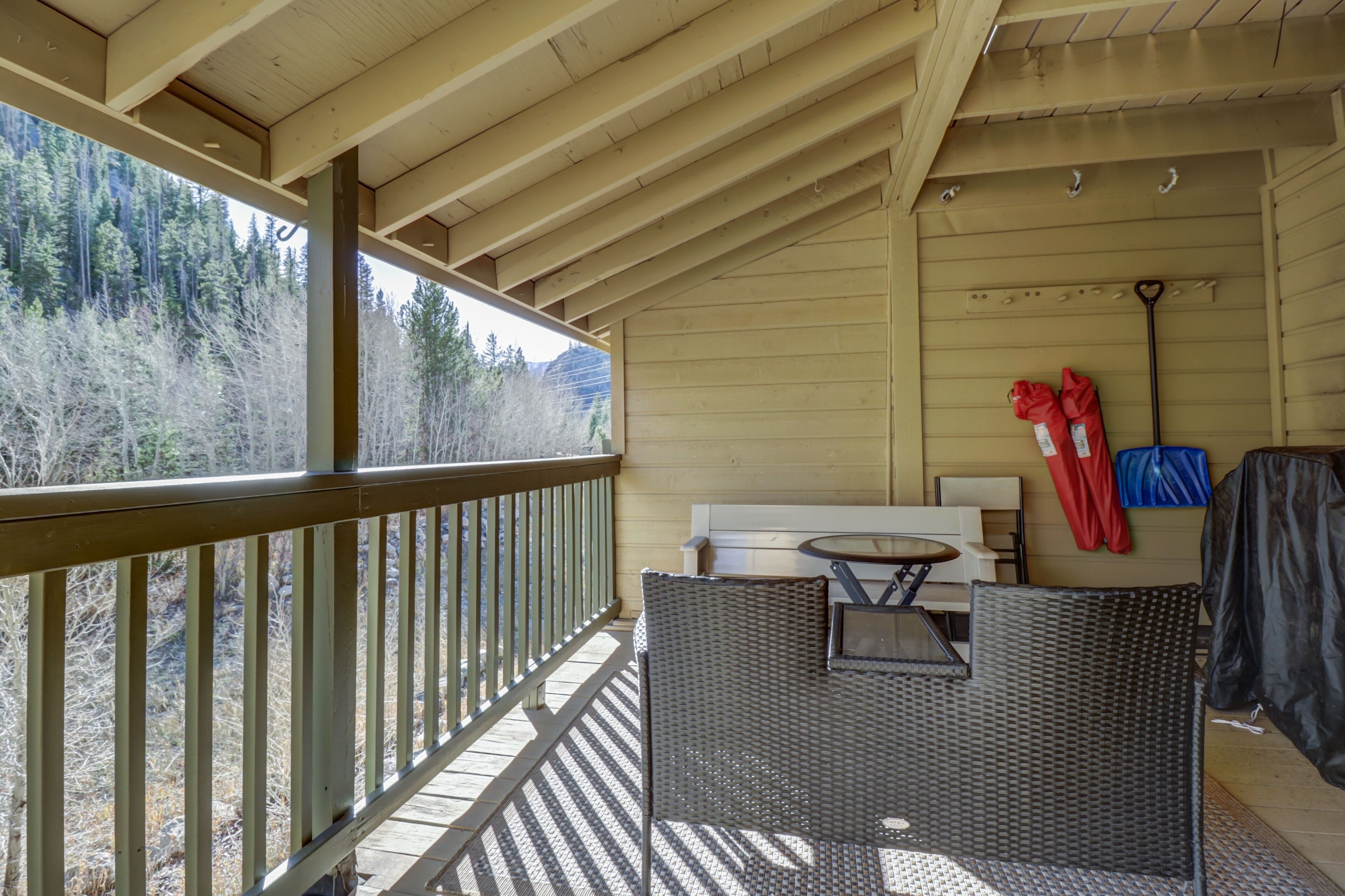 A covered outdoor area with a wooden deck, wicker furniture, and a view of a forest.