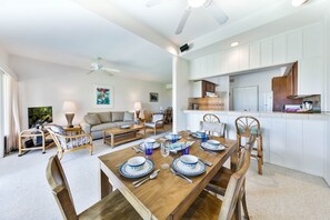 Amplia sala de estar con una mesa de comedor lista para seis personas, rodeada de asientos cómodos. Se ve una cocina con armarios blancos al fondo.