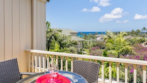A balcony with wicker chairs, a table with a red tray, and a glass of wine. The view includes a pool, palm trees, and a clear blue sky.
