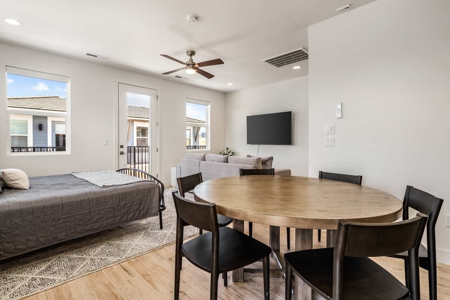 A modern room with a bed, a dining table with chairs, a ceiling fan, and a TV mounted on the wall.