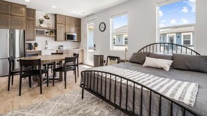 Quarto moderno com estrutura de cama de metal, uma janela grande com vista para um edifício e área de cozinha com armários de madeira e micro-ondas.