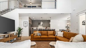 A spacious living area with a large flat-screen TV, a brown sofa, and two white armchairs. A kitchen with modern appliances and a dining area with a table and chairs are visible in the background. - Desert Brooke Villa 6 Bedroom Townhouse (St. George)