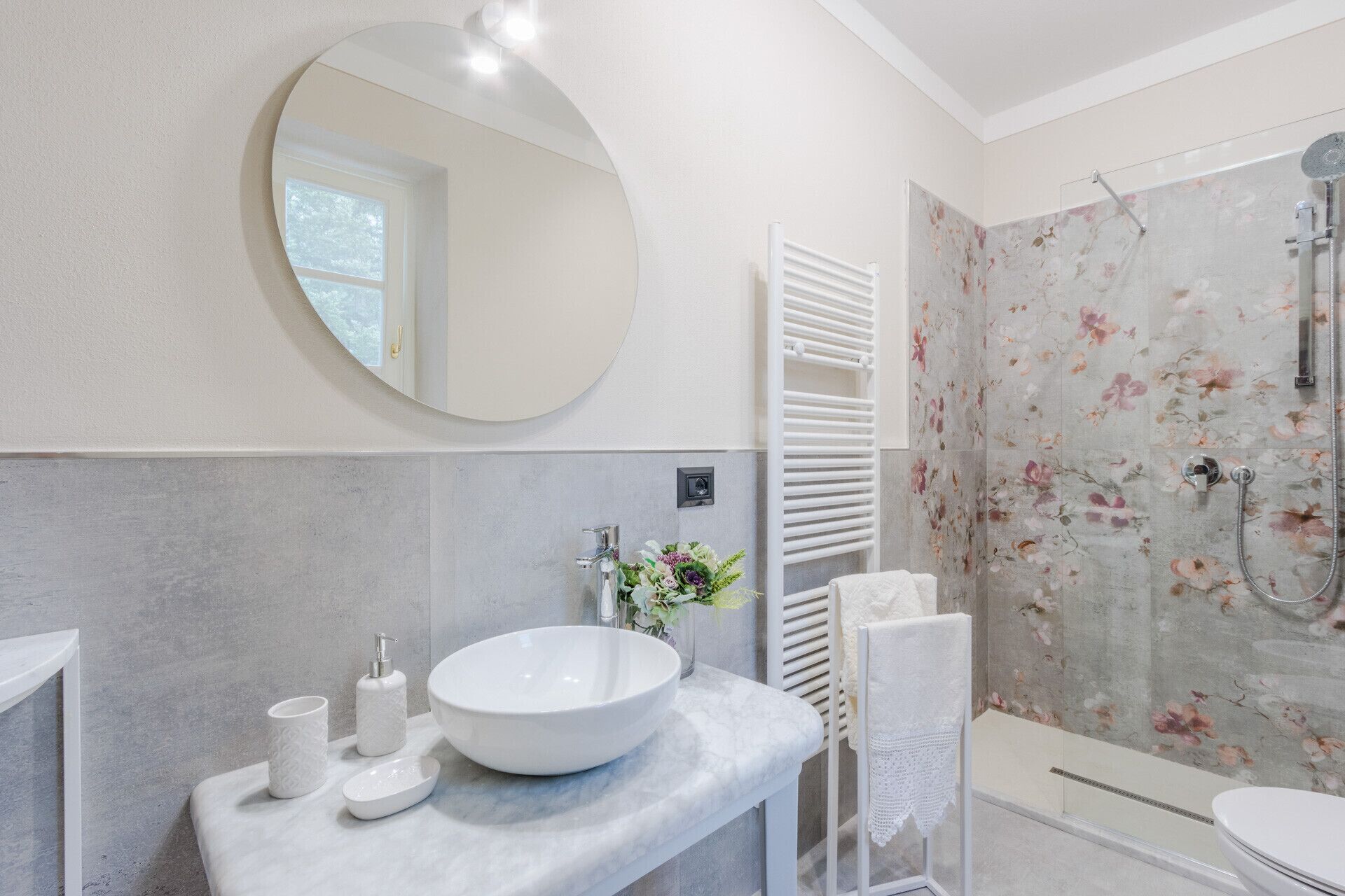 Un baño moderno con un espejo redondo, un lavamanos grande de color blanco, una ducha con azulejos florales y un perchero para toallas.