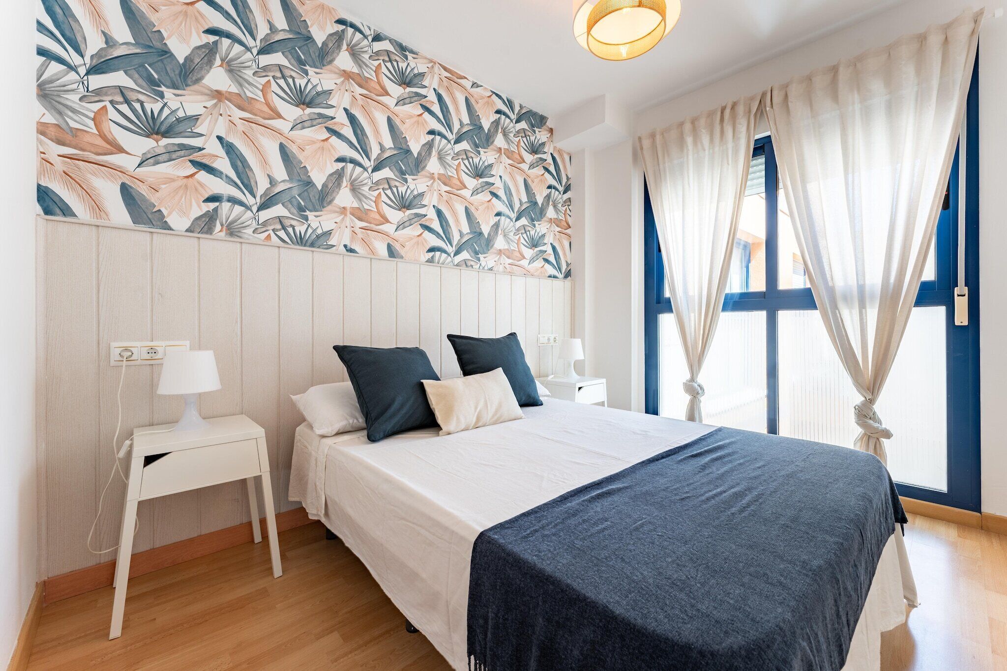 Een slaapkamer met een bed, nachtkastjes, een muur met een tropisch bladmotief en een raam met gordijnen.