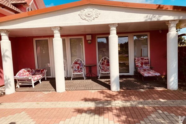 Une terrasse couverte avec des colonnes blanches, un mur rouge et deux espaces de détente en plein air agrémentés de coussins à motifs floraux.