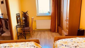 Une chambre avec deux lits, une armoire en bois, un radiateur, une fenêtre avec des rideaux et un bureau avec une chaise.