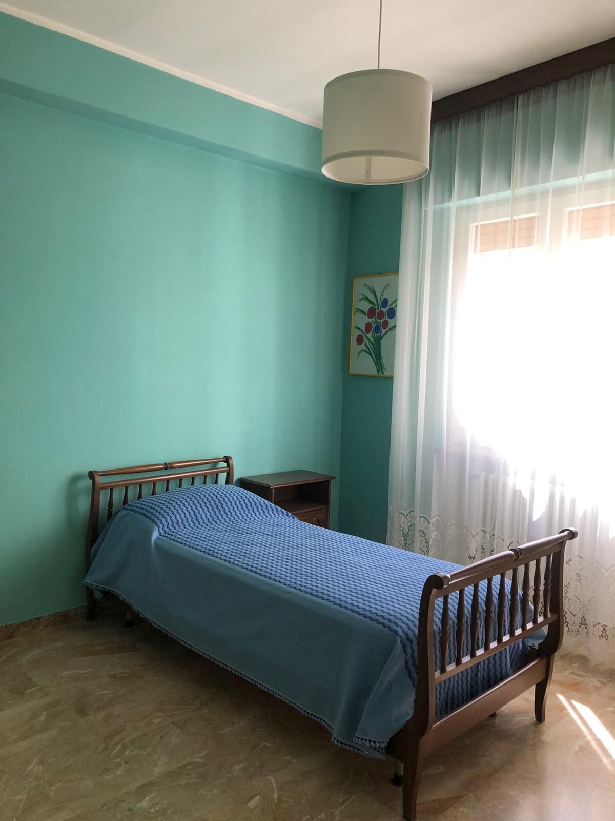 A bedroom with a single bed, a nightstand, a hanging light fixture, and a window with curtains.