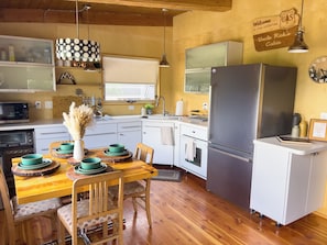 A compact kitchen with a refrigerator, microwave, and oven. There is a dining table with chairs and a vase of dried flowers. The kitchen has wooden floors and a window with a view.