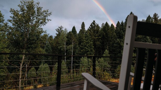 Una terraza de madera con barandilla, con vista a un área boscosa y un arcoíris en el cielo.