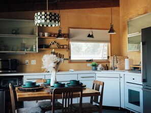 A kitchen with wooden cabinets, a dining table with chairs, a hanging light fixture, and a window with blinds. - Pike National Forest Cabin with Mountain Views, Deck & Wood Fireplace (Woodland Park)
