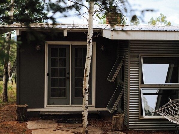 A small, single-story building with a front door and windows, surrounded by trees and a wooden post.