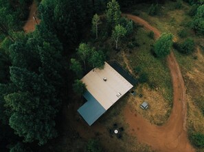 Vista aérea de una casa de un piso con techo plano, rodeada de un bosque denso. Un camino de tierra lleva a la casa y hay un pequeño claro con una mesa y sillas.