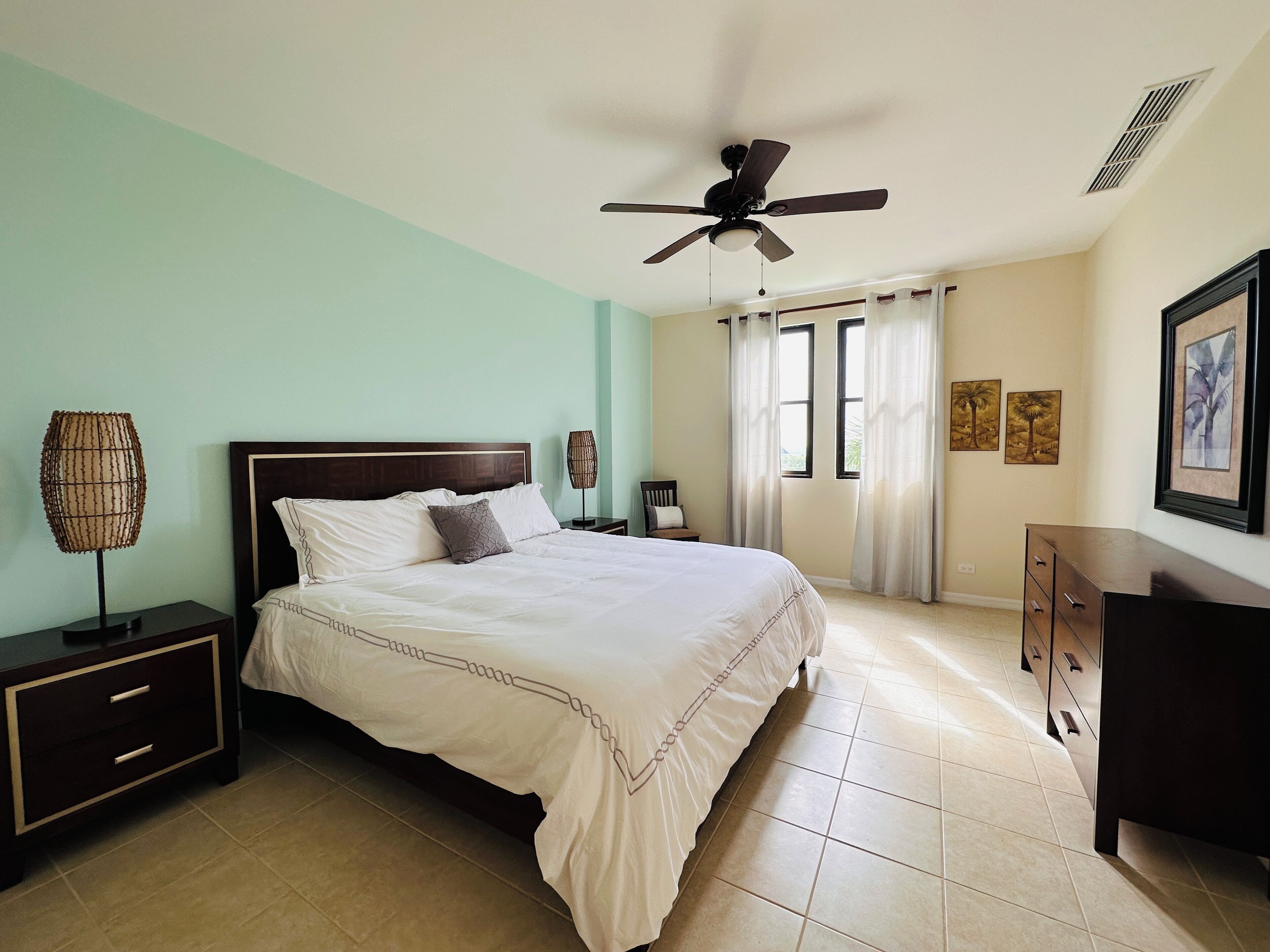 A bedroom with a bed, nightstands, a dresser, a ceiling fan, and a window with curtains.