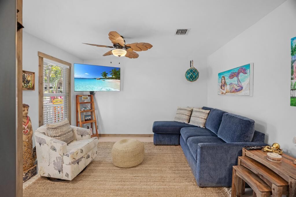 A living room with a blue sofa, a white armchair, a ceiling fan, and a TV displaying a beach scene.