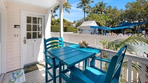 Eine Veranda mit einer weißen Tür, einem blauen Tisch und Stühlen sowie im Hintergrund Blick auf Palmen und einen Strand.