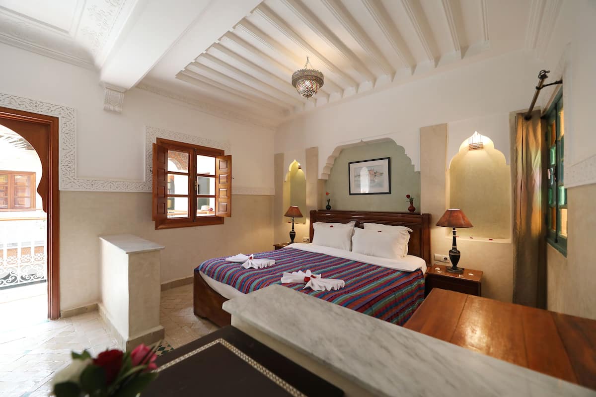 Une chambre d’hôtel avec un grand lit, une table de chevet en bois, un bureau, une chaise, une petite table, un mur décoratif et une fenêtre avec des rideaux.