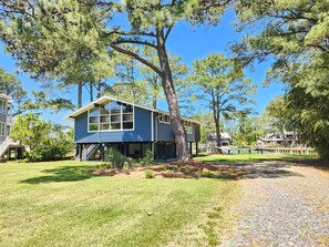 A two-story house with a large tree in front, a grassy yard, and a dock extending into a body of water.