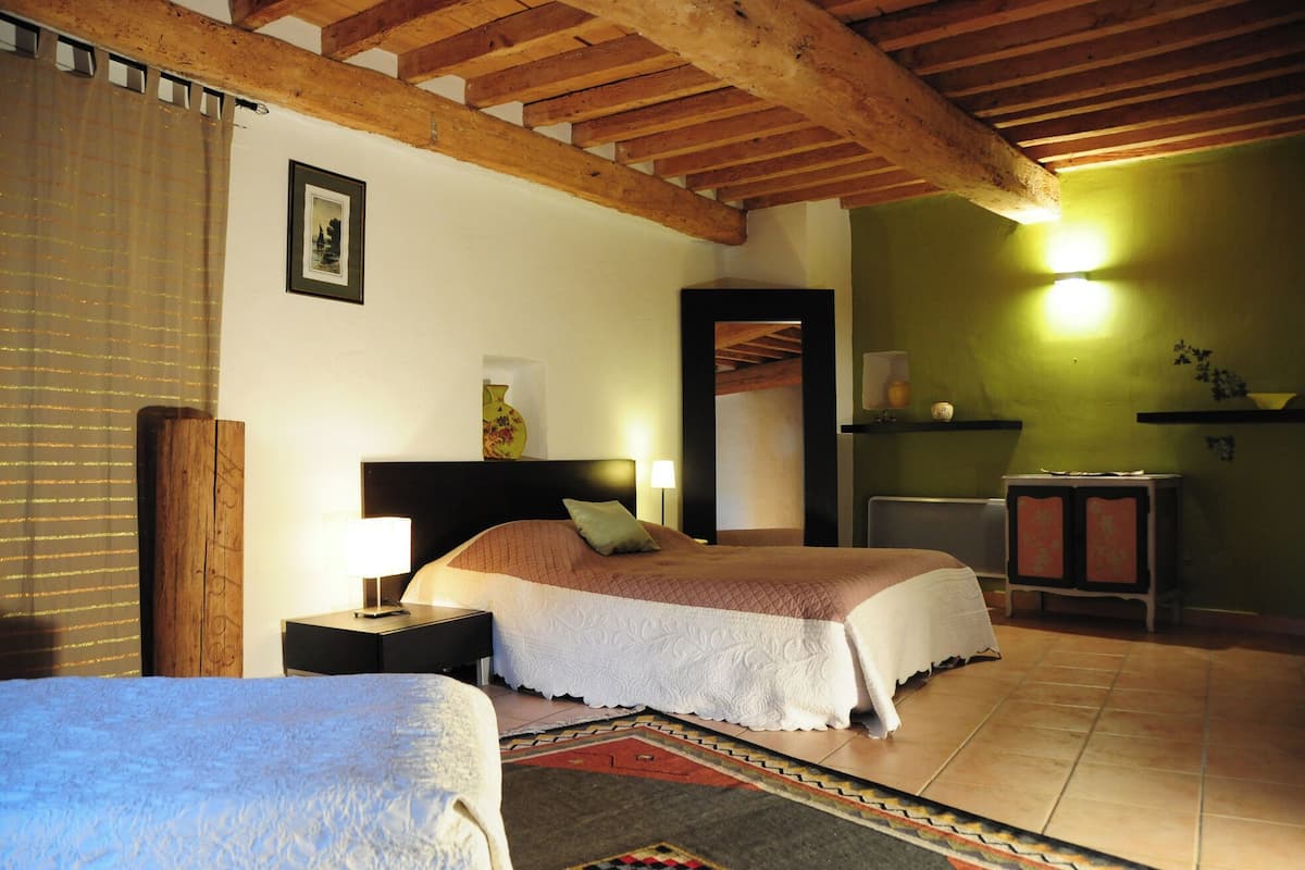 Quarto com teto de madeira, cama com colcha branca, mesa de cabeceira com abajur, pequena mesa com um vaso e parede verde com luminária.