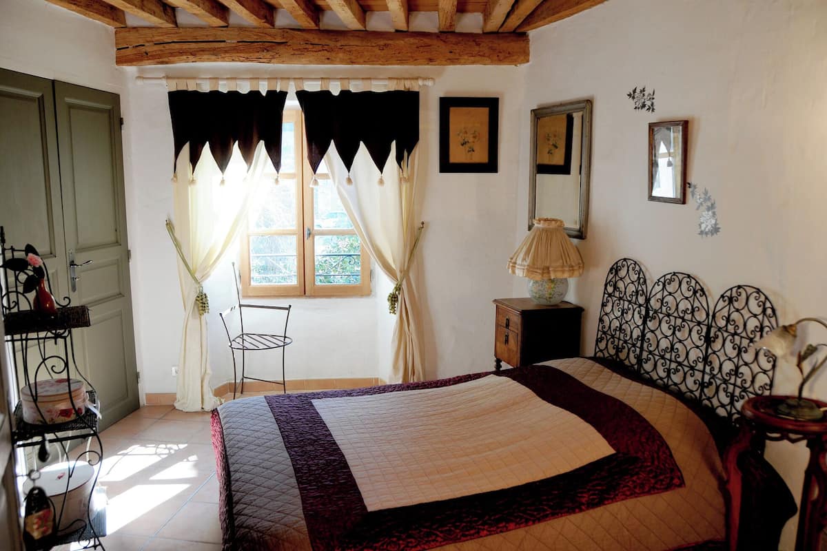 Quarto com cama, janela com cortinas, mesa de cabeceira com abajur e uma cadeira.