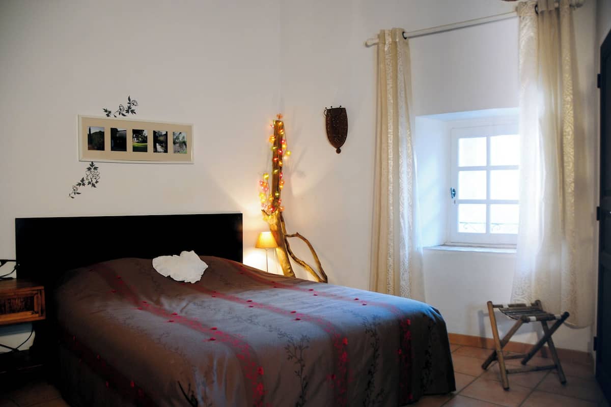 Quarto com cama, uma mesinha, uma cadeira, janela com cortinas e detalhes decorativos na parede, incluindo um porta-retrato e um enfeite fixado.