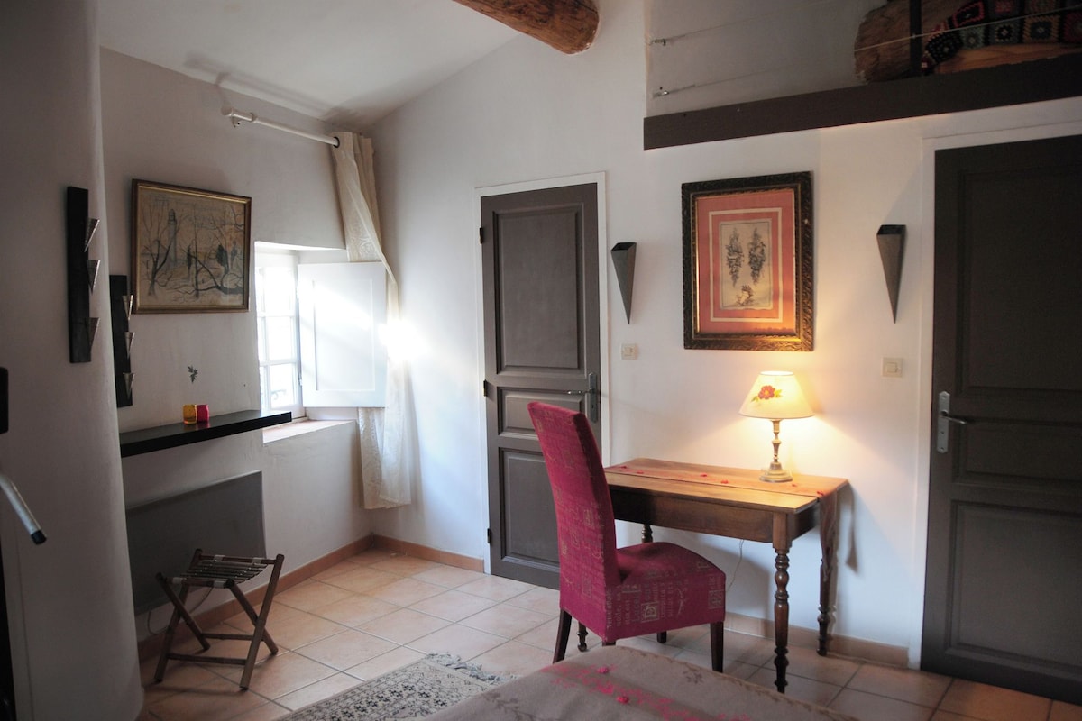 A small, well-lit room with a desk, chair, and a lamp. There are two doors, one with a window, and a framed picture on the wall.