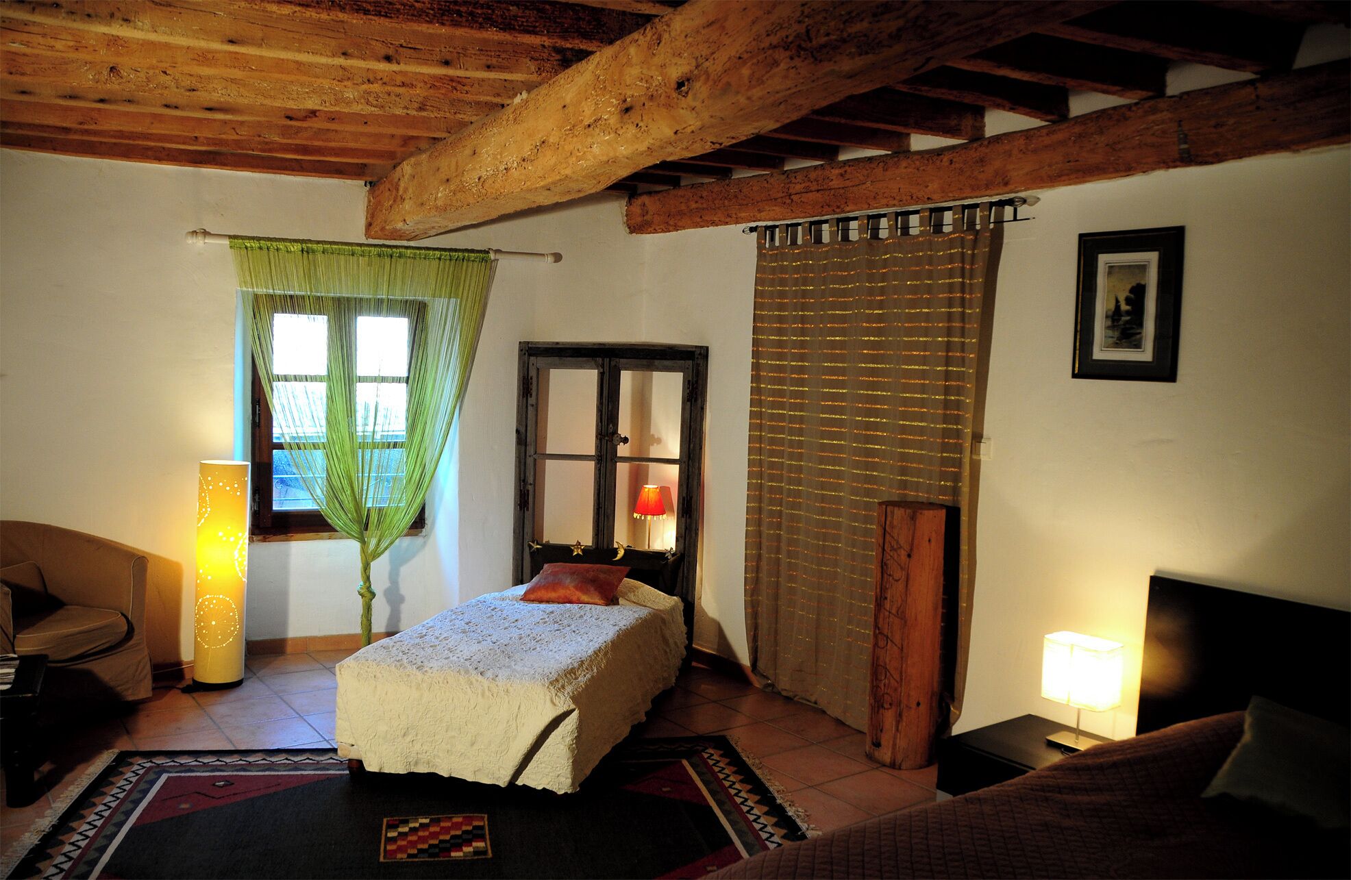 Ein gemütliches Zimmer mit Bett, Sessel, Tisch und Lampe. Ein Fenster mit Vorhängen und eine Tür mit Spiegel.