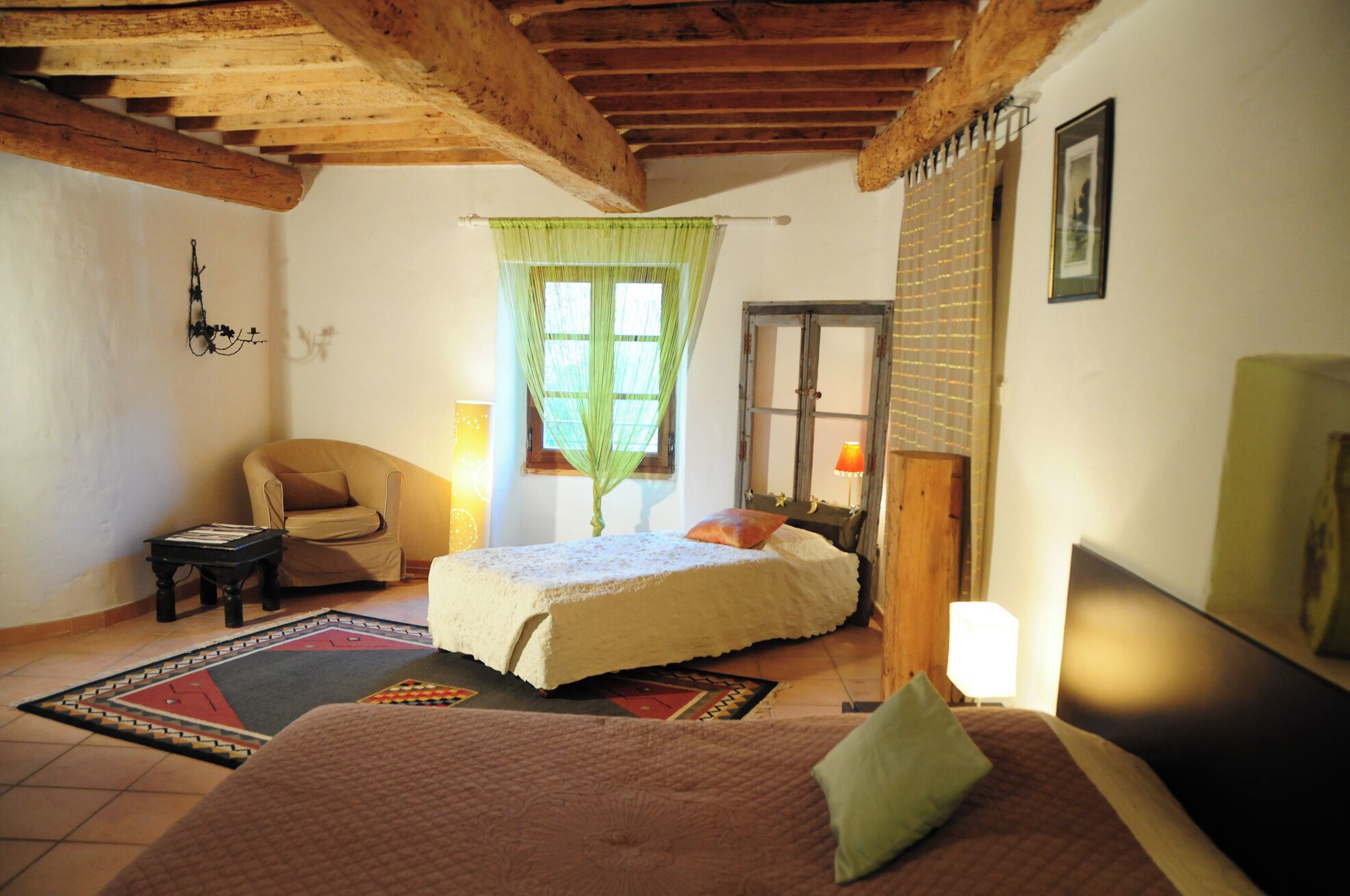 Een gezellige slaapkamer met een groot bed, een kleiner bed, een stoel en een bureau. Er is een raam met groene gordijnen en een lamp op het bureau.