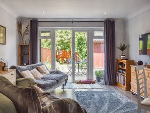 Ett vardagsrum med en soffa, ett soffbord, en bokhylla och en tv. Det finns en skjutdörr i glas som leder ut till en uteplats med ett bord och stolar.