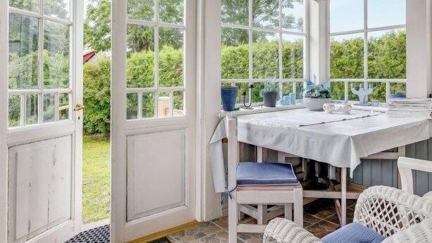 Et orangeri med et bord, stole, store vinduer og potteplanter.