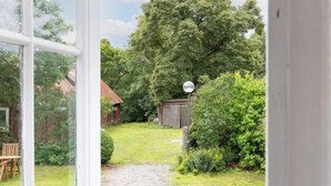 Udsigt gennem en dør til en baghave med en grussti, en træbænk og en rød bygning i baggrunden.