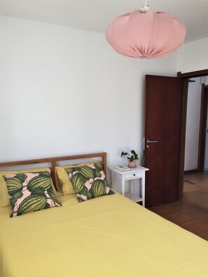 A bedroom with a bed, two pillows, a bedside table with a book and a plant, a wooden door, and a pink hanging light fixture. - Casa Capuchos. Close to Lisbon with Ocean View (Caparica)