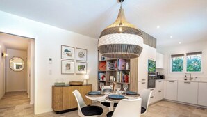 Een moderne keuken met witte kasten, een eethoek met witte stoelen, een grote geweven hanglamp en een boekenkast vol boeken en decoratieve objecten.