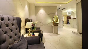 A modern hotel lobby with a tufted armchair, a table with a lamp and vase, and a decorative wall piece. - A Boutique Hotel View (New Delhi)