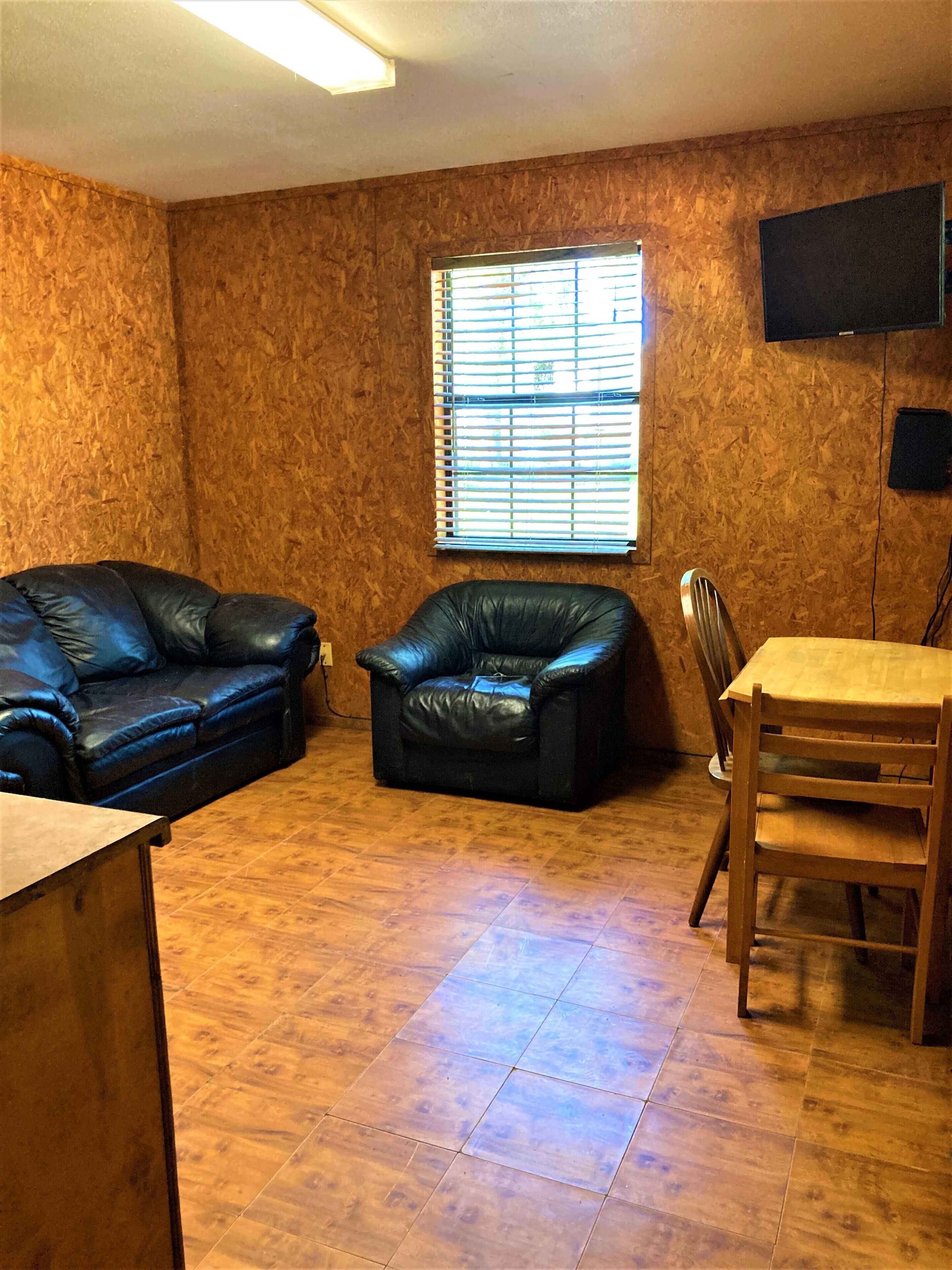 Une pièce avec un plancher en bois, un canapé en cuir noir, un fauteuil en cuir noir, une table à manger en bois avec des chaises, et une fenêtre avec des stores.