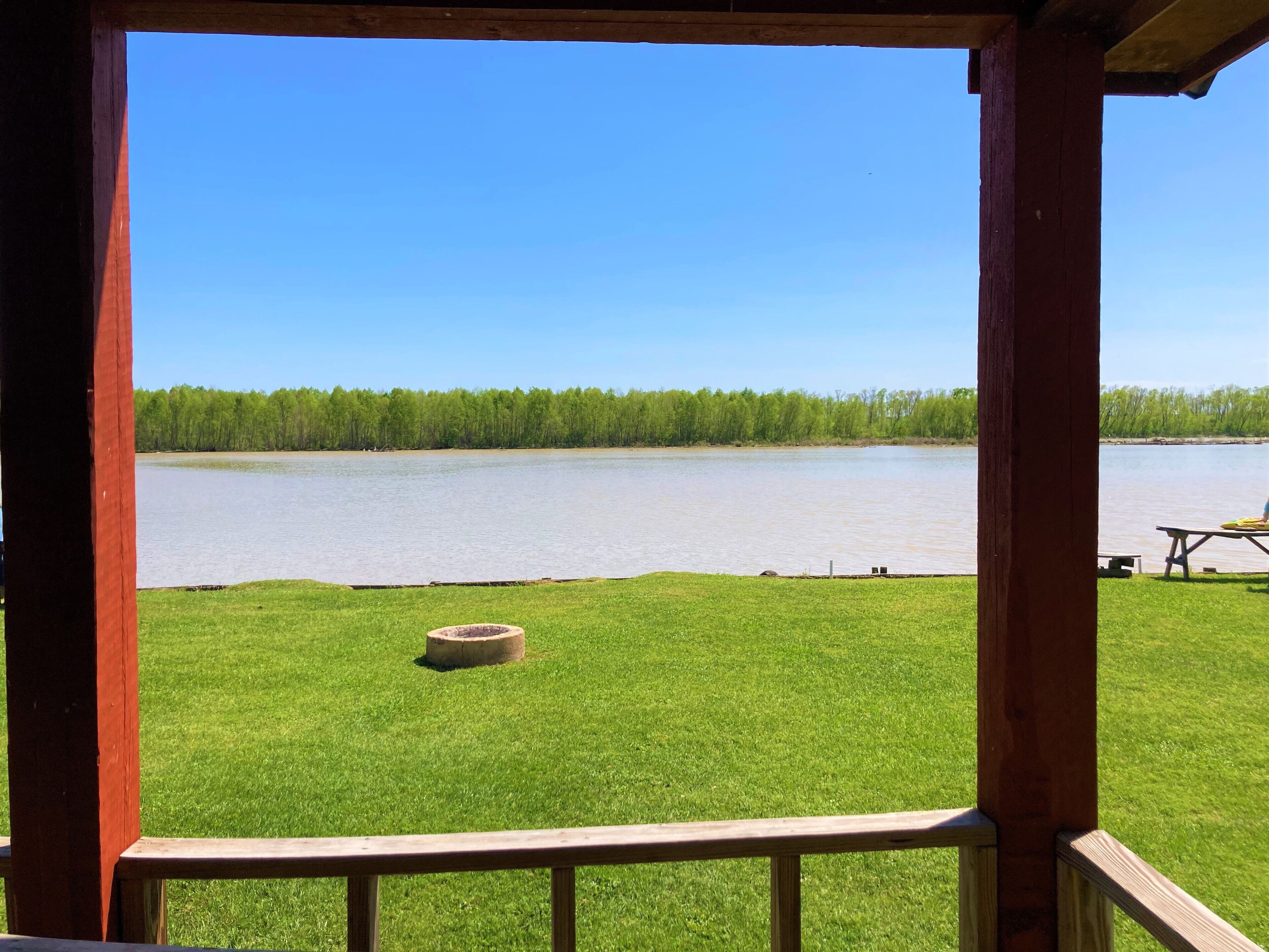 A view from a wooden deck overlooking a calm lake, with a grassy area and a picnic table in the foreground.
