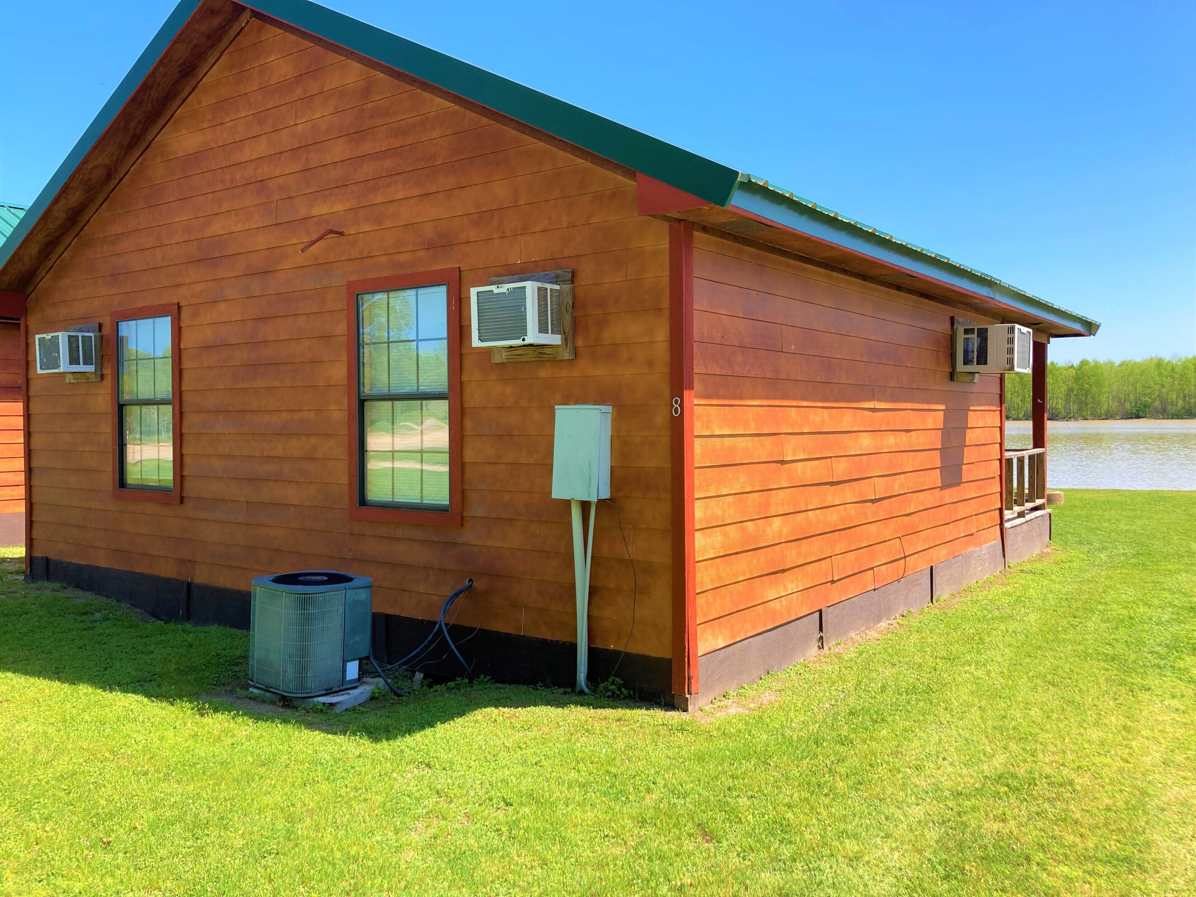 A wooden cabin with a green roof, air conditioning units, and a small outdoor area with grass.