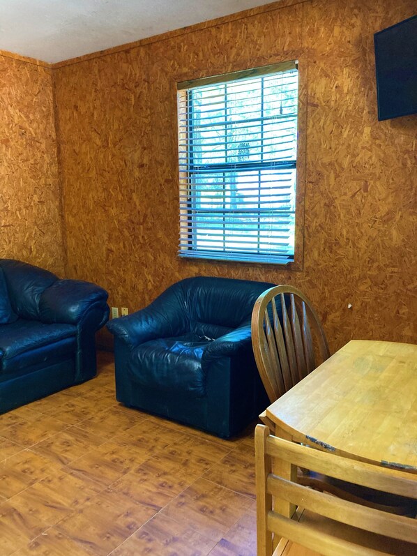 A room with a wooden wall, a window with blinds, a blue leather sofa, a wooden dining table with chairs, and a wooden chair. - 1075 Marina Dr (Trinity)