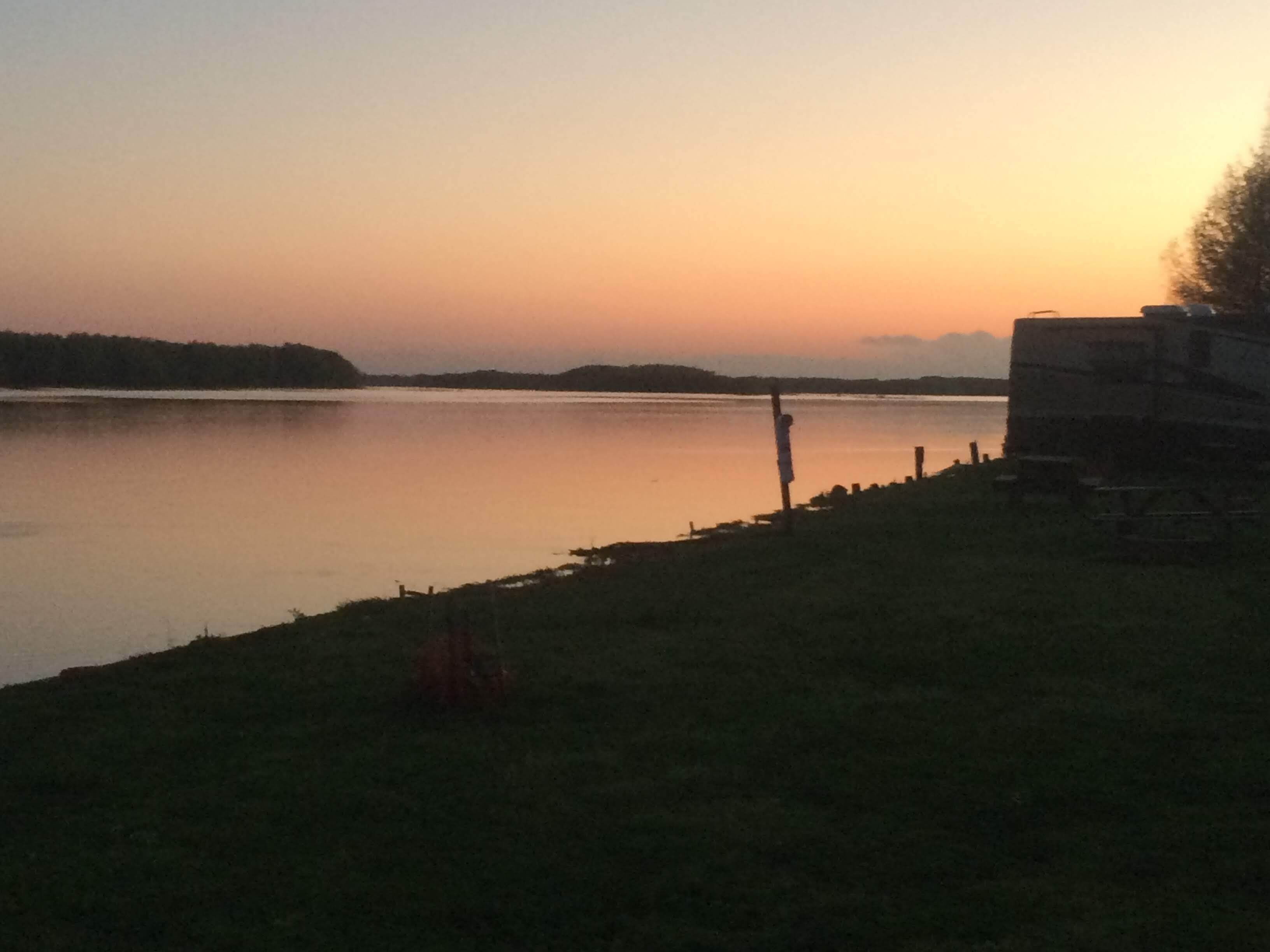 Un lac paisible au coucher du soleil, avec une aire de camping et une caravane sur la droite.