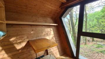 Eine Holzhütte mit geneigtem Dach, einem kleinen Tisch, einem Stuhl und einem großen Fenster mit Blick auf einen Wald.
