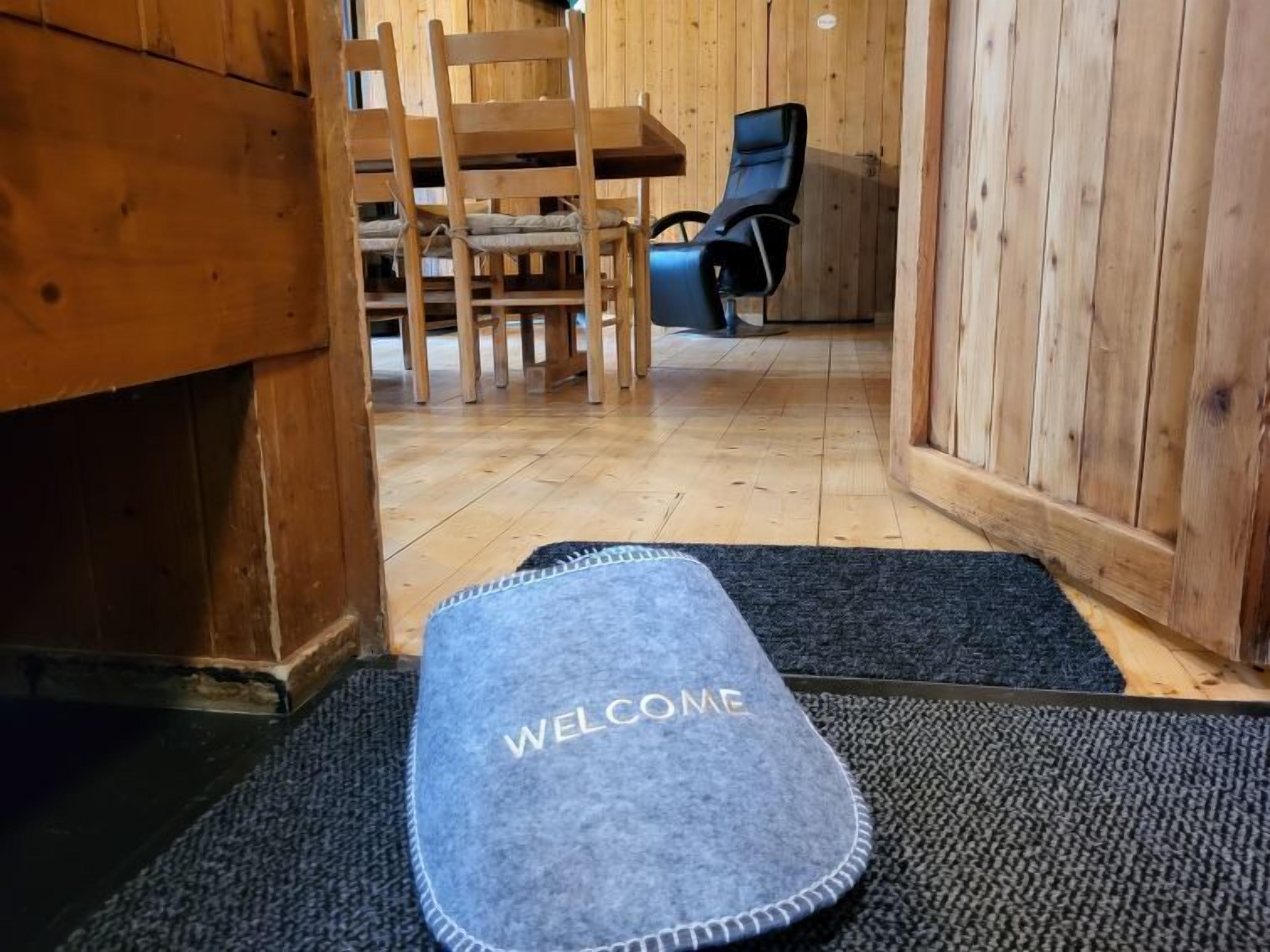 Een houten interieur met een 'WELCOME' mat bij de ingang, houten stoelen en een zwarte stoel.