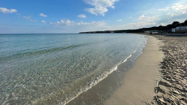 A beach with clear water, a sandy shore, and distant buildings. - Acquamarina (Melendugno)