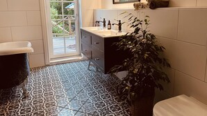 Un bagno con lavandino bianco, mobile contenitore scuro, piastrelle decorative a pavimento, una pianta in vaso e una finestra vista verde.