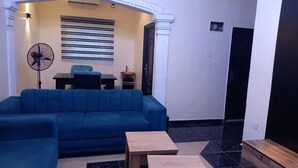 Un salón con un sofá azul, dos mesitas de madera, un tapiz blanco y negro, y un ventilador.