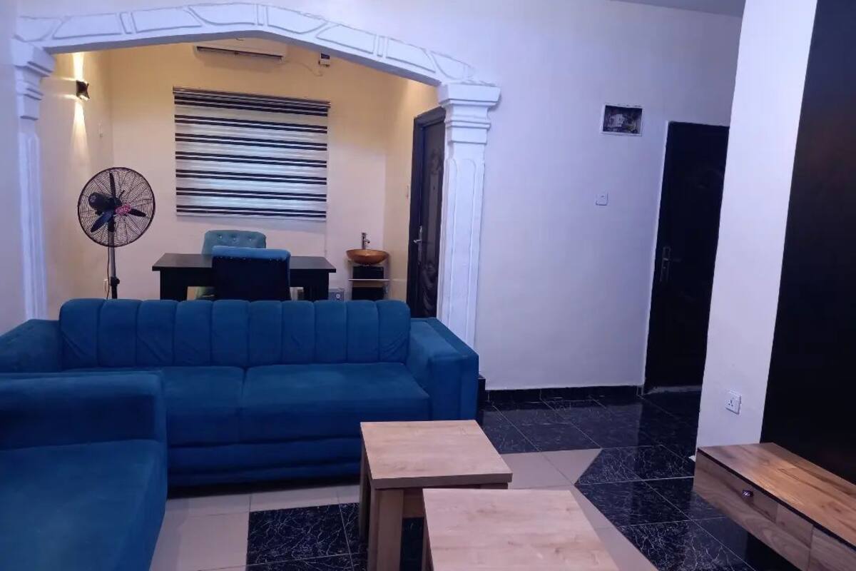 A living room with a blue sofa, two wooden side tables, a black and white striped wall hanging, and a fan.